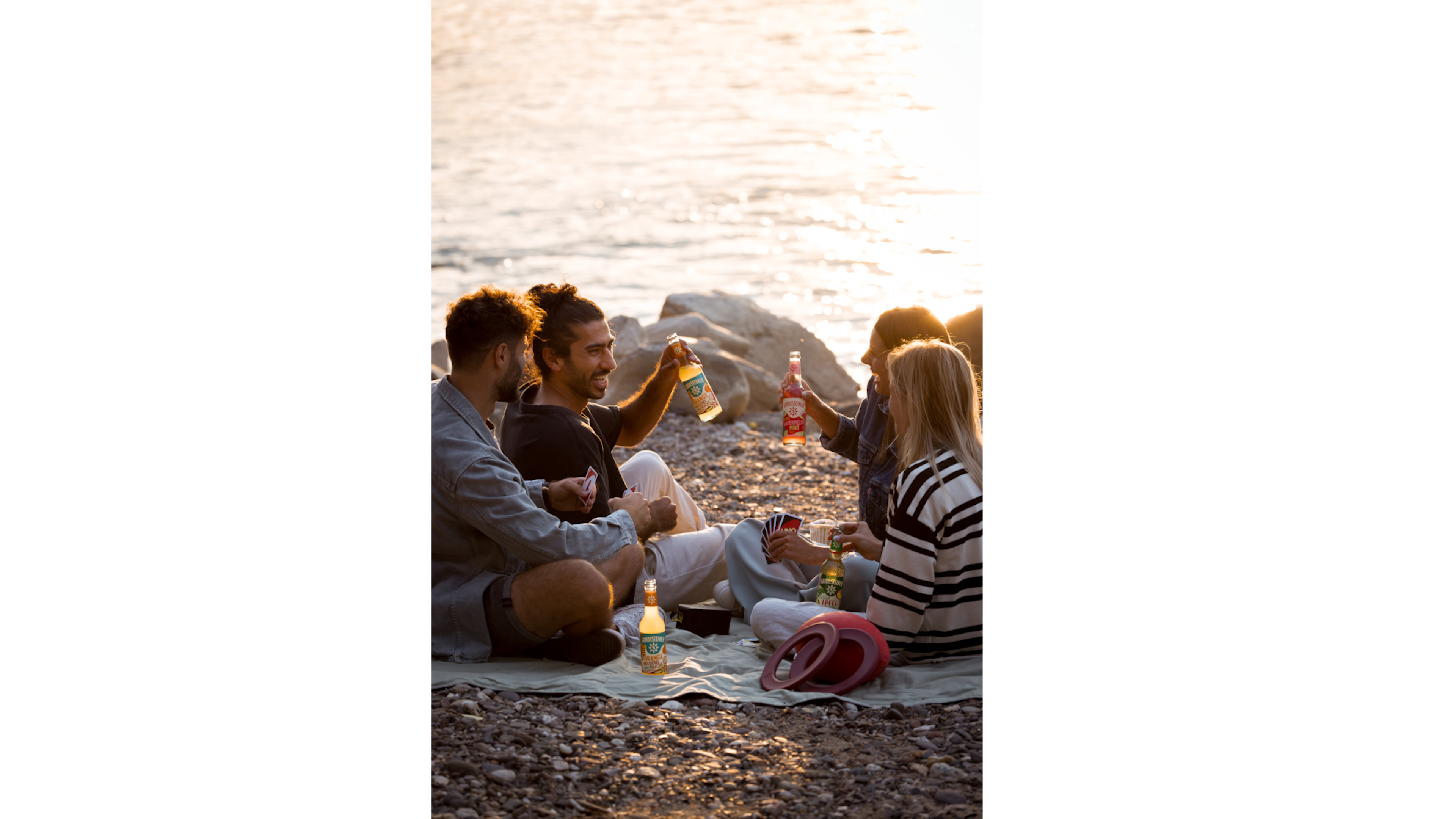 Eine Gruppe junger Leute sitzt entspannt beim Sonnenuntergang am Fluss und trinkt Gerolsteiner Erfrischungsgetränke in der kleinen Glasflasche
