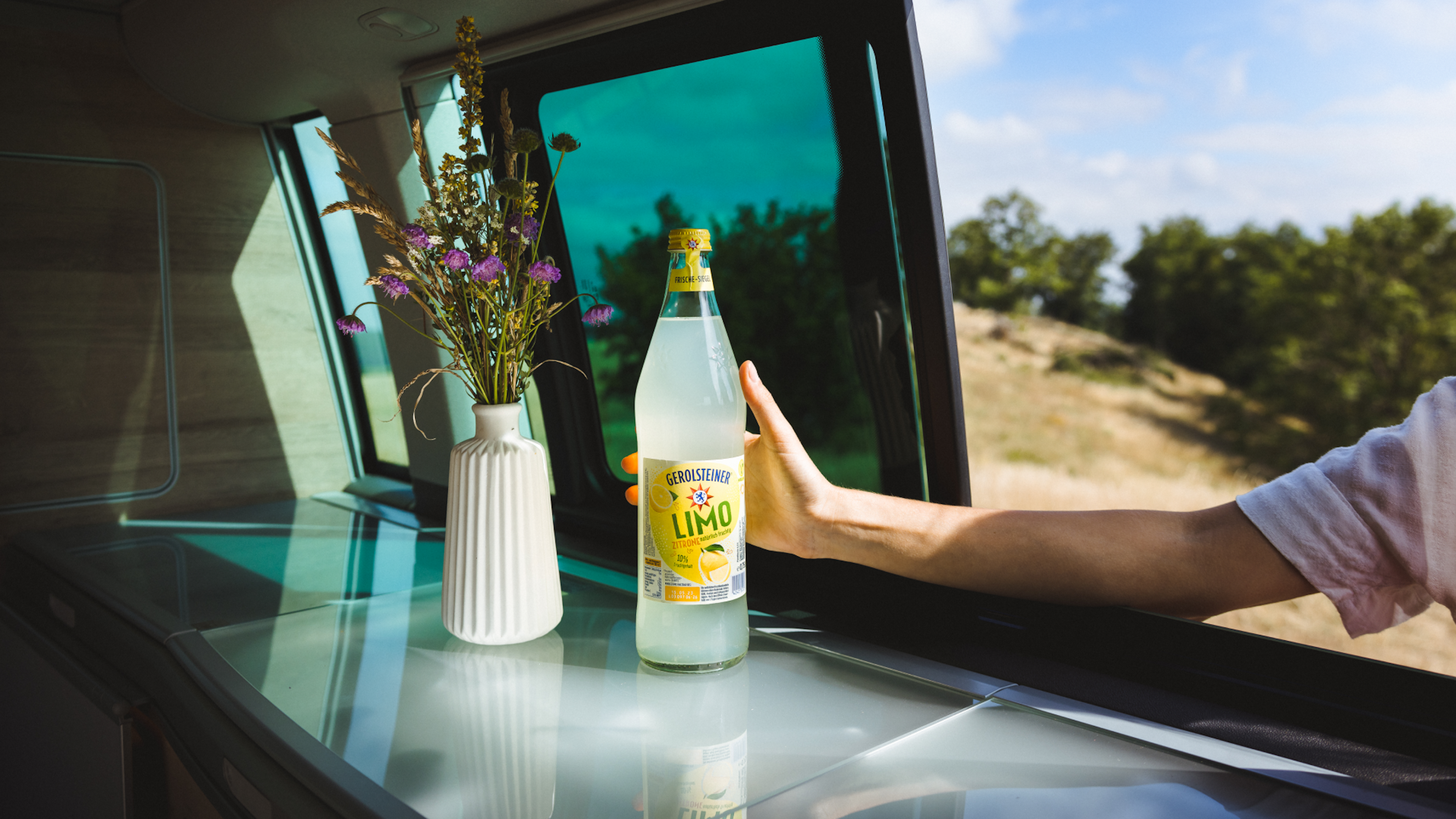 Eine Flasche Zitronenlimonade steht vor einer geöffneten Wagentür neben einer Vase mit Wildblumen.