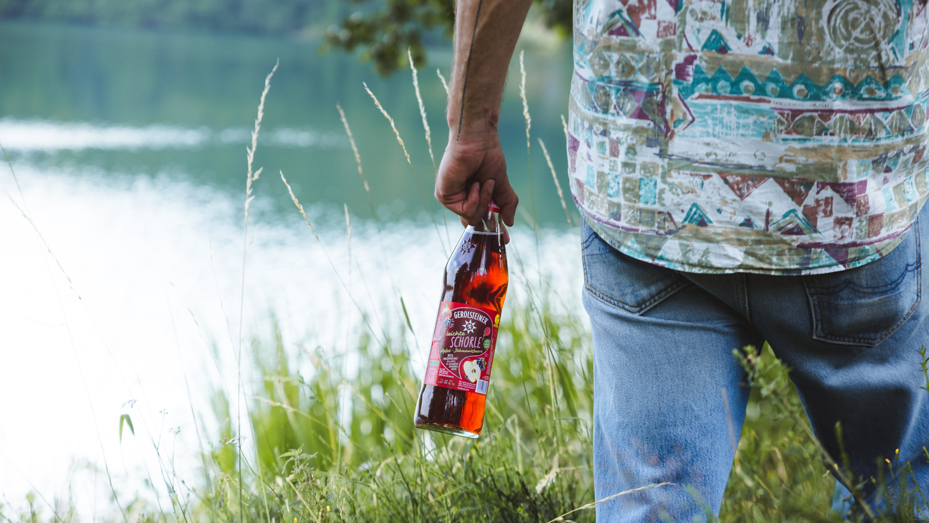 Eine Person hält eine Flasche Apfel-Johannisbeere-Schorle neben einem See.