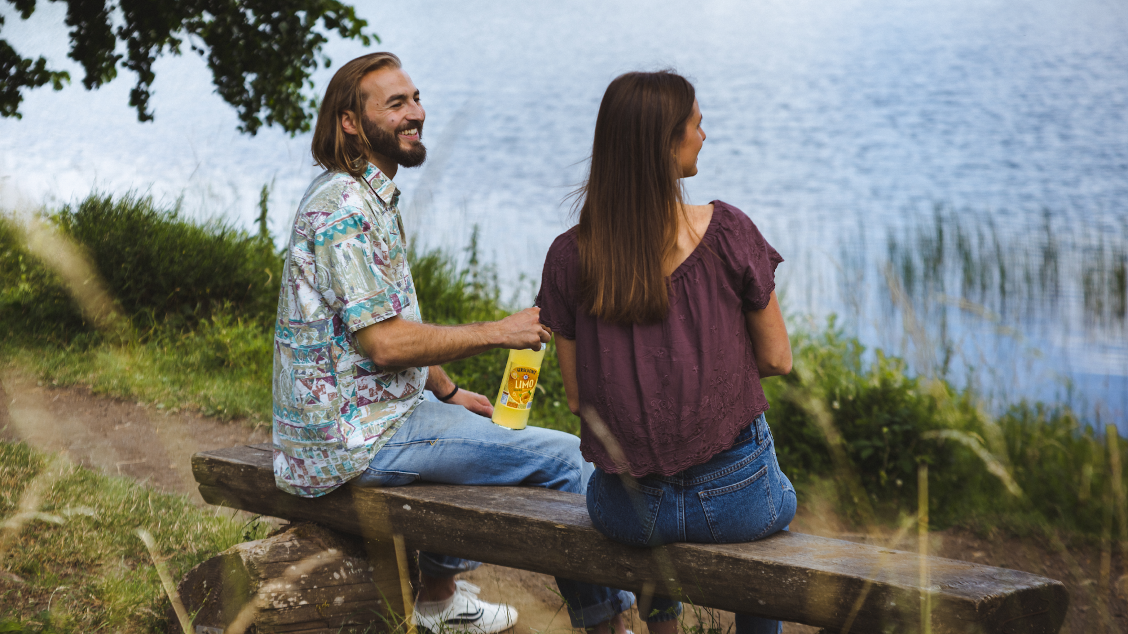 Ein Mann und eine Frau sitzen auf einer Bank am See und haben eine Flasche Orangenlimonade dabei