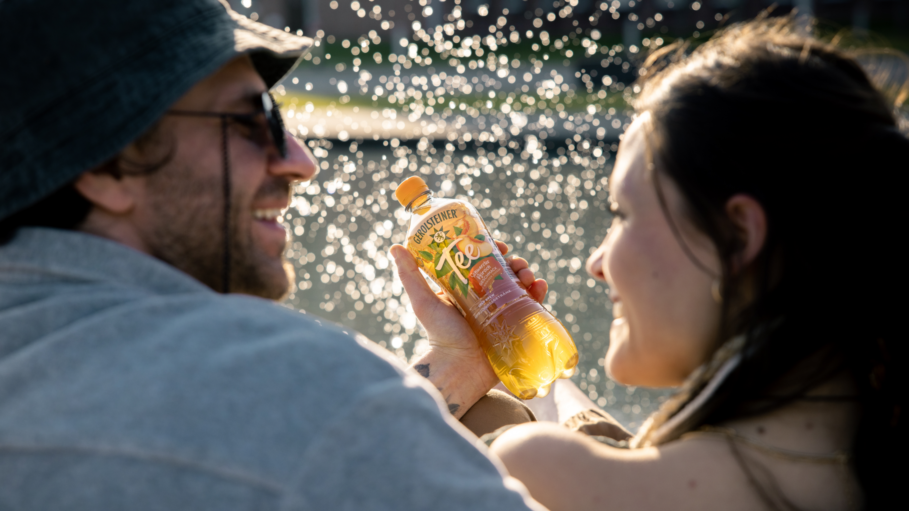 Ein Paar hält sich lächelnd eine Flasche Eistee in der Sonne entgegen.