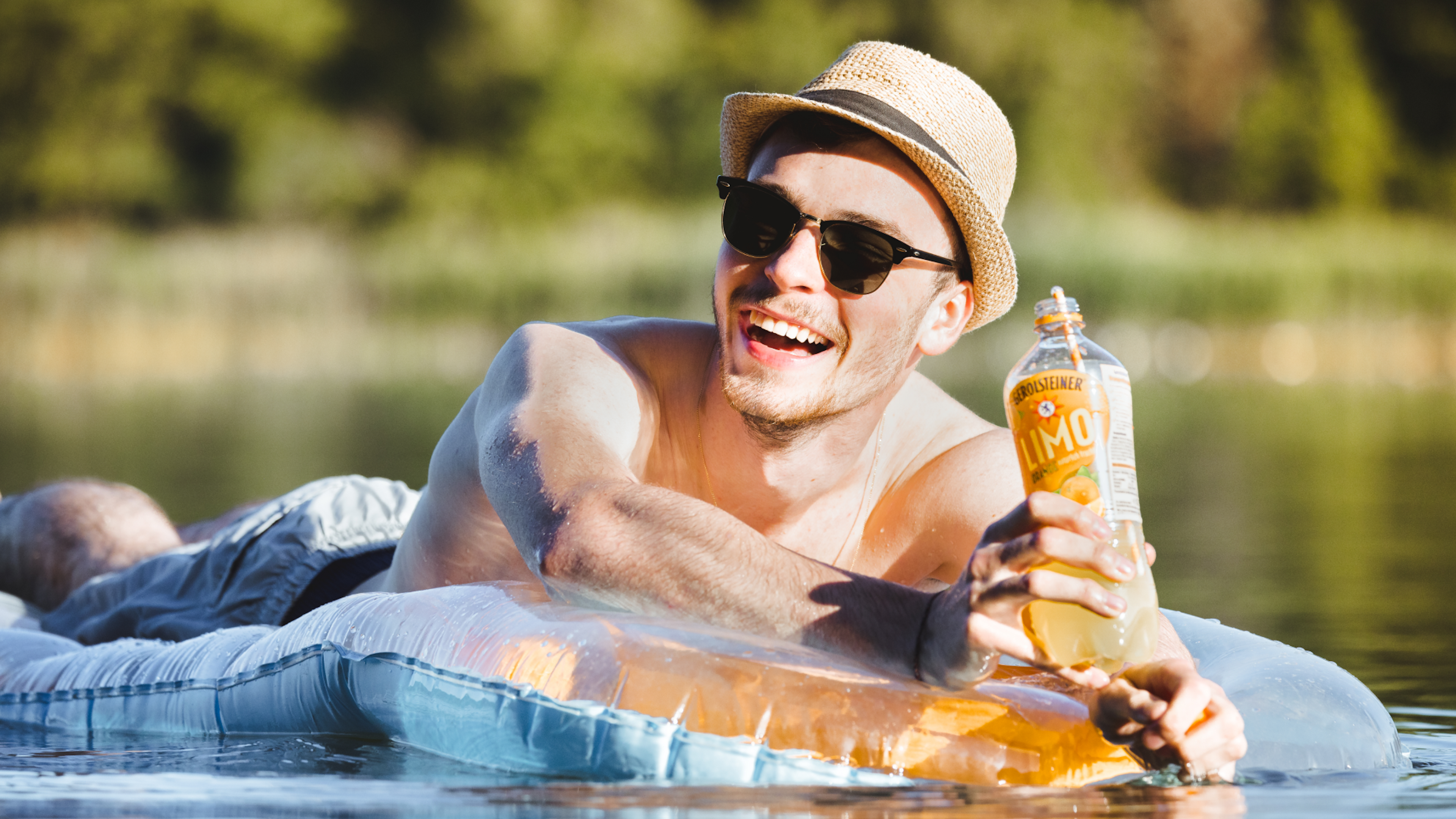 Ein lächelnder Mann auf einem Schwimmreifen im Wasser hält eine Flasche Leichte Limonade.