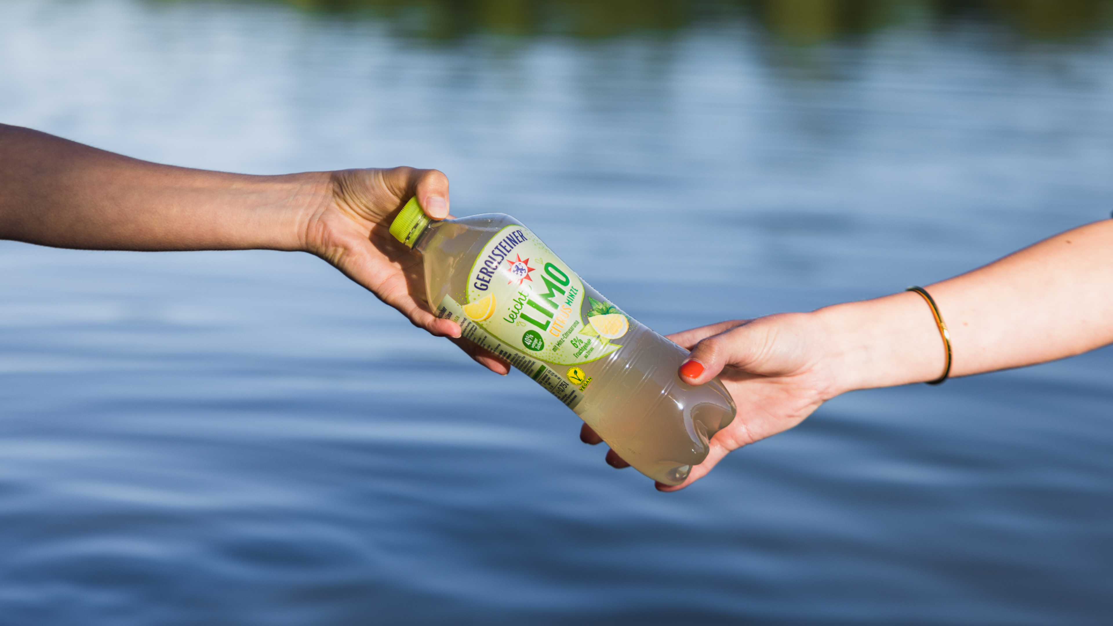 Zwei Hände tauschen eine Flasche Limonade auf einem See aus.