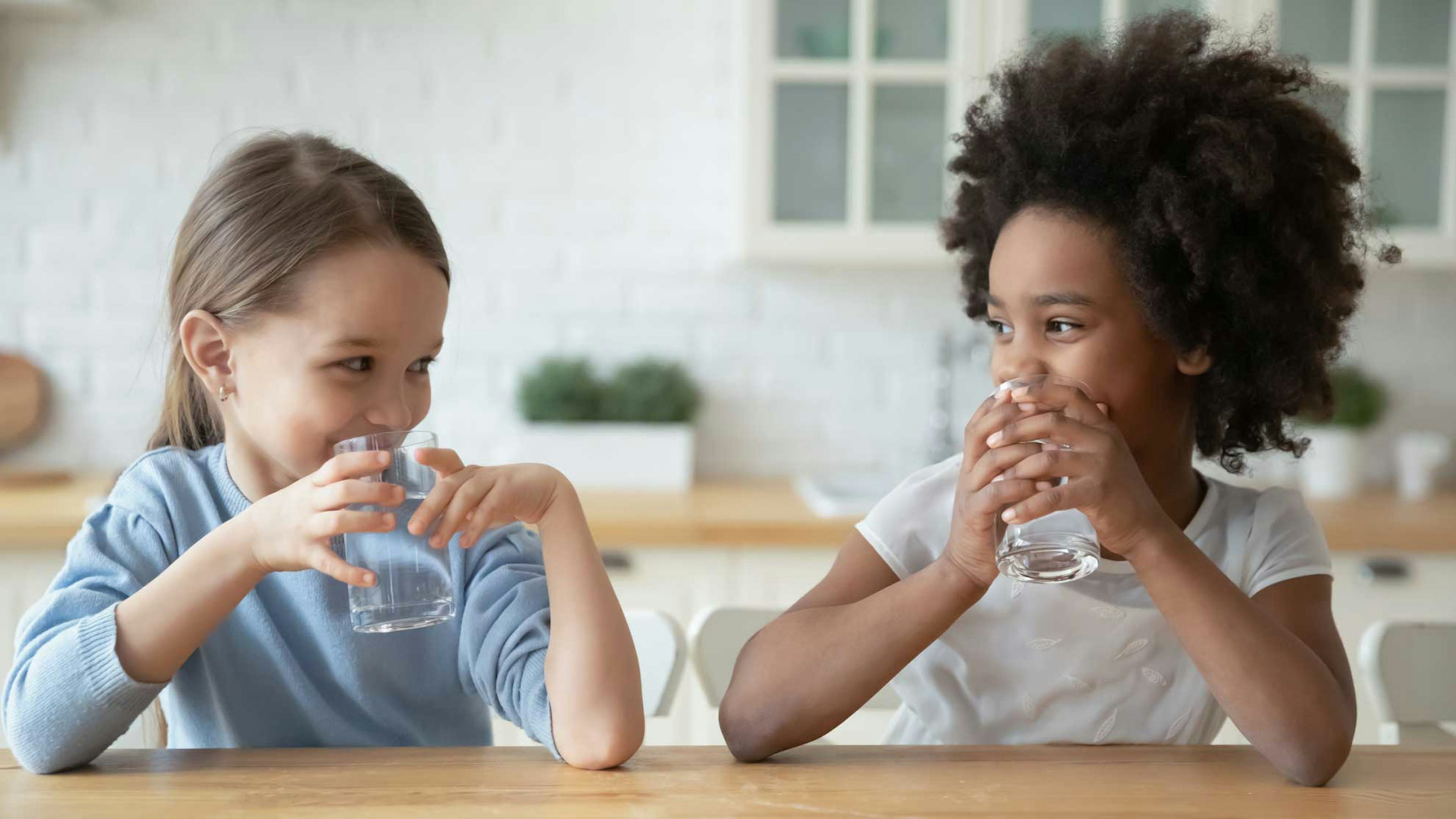 Zwei junge Mädchen sitzen an einem Holztisch und trinken je ein Glas Wasser