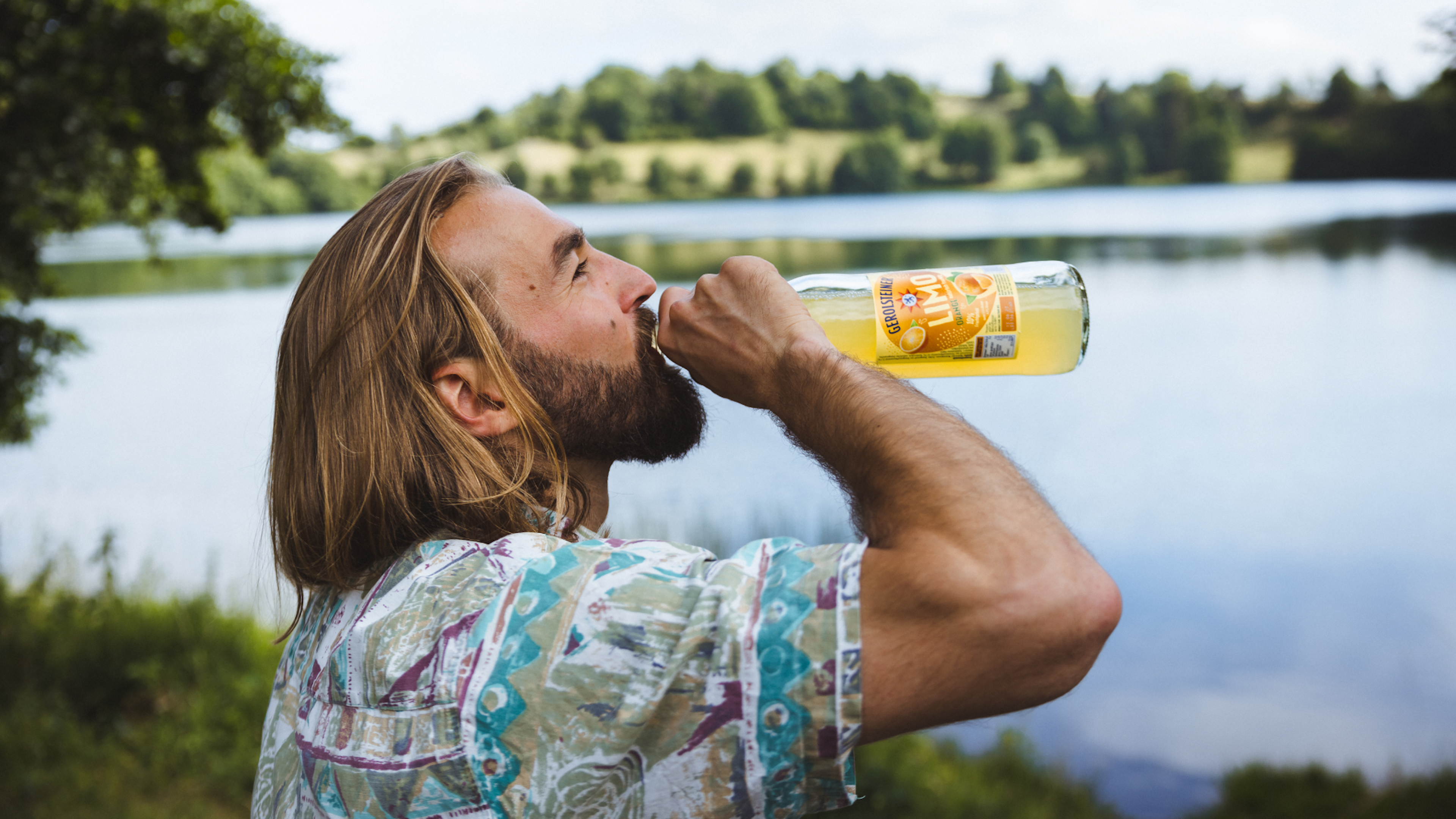 Ein Mann in Sommerkleidung trinkt eine Flasche Orangenlimonade in der Natur.
