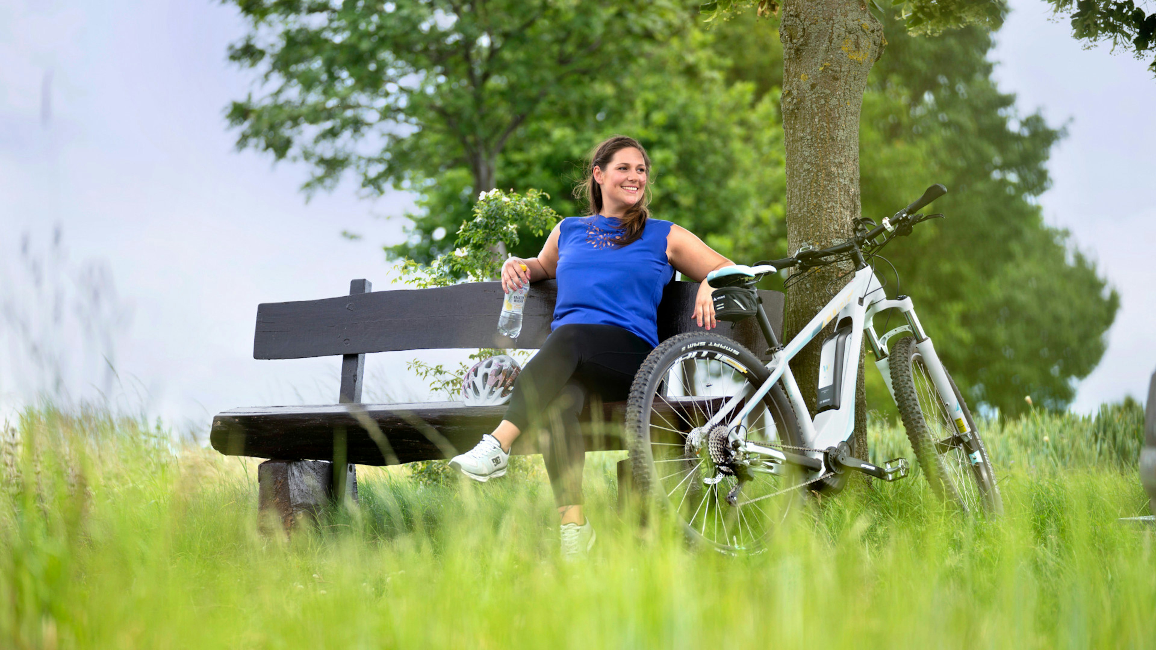 Eine Frau auf einer Bank im Freien mit einer Flasche Gerolsteiner Kräuterwasser in der Hand, ein Fahrrad steht neben ihr.