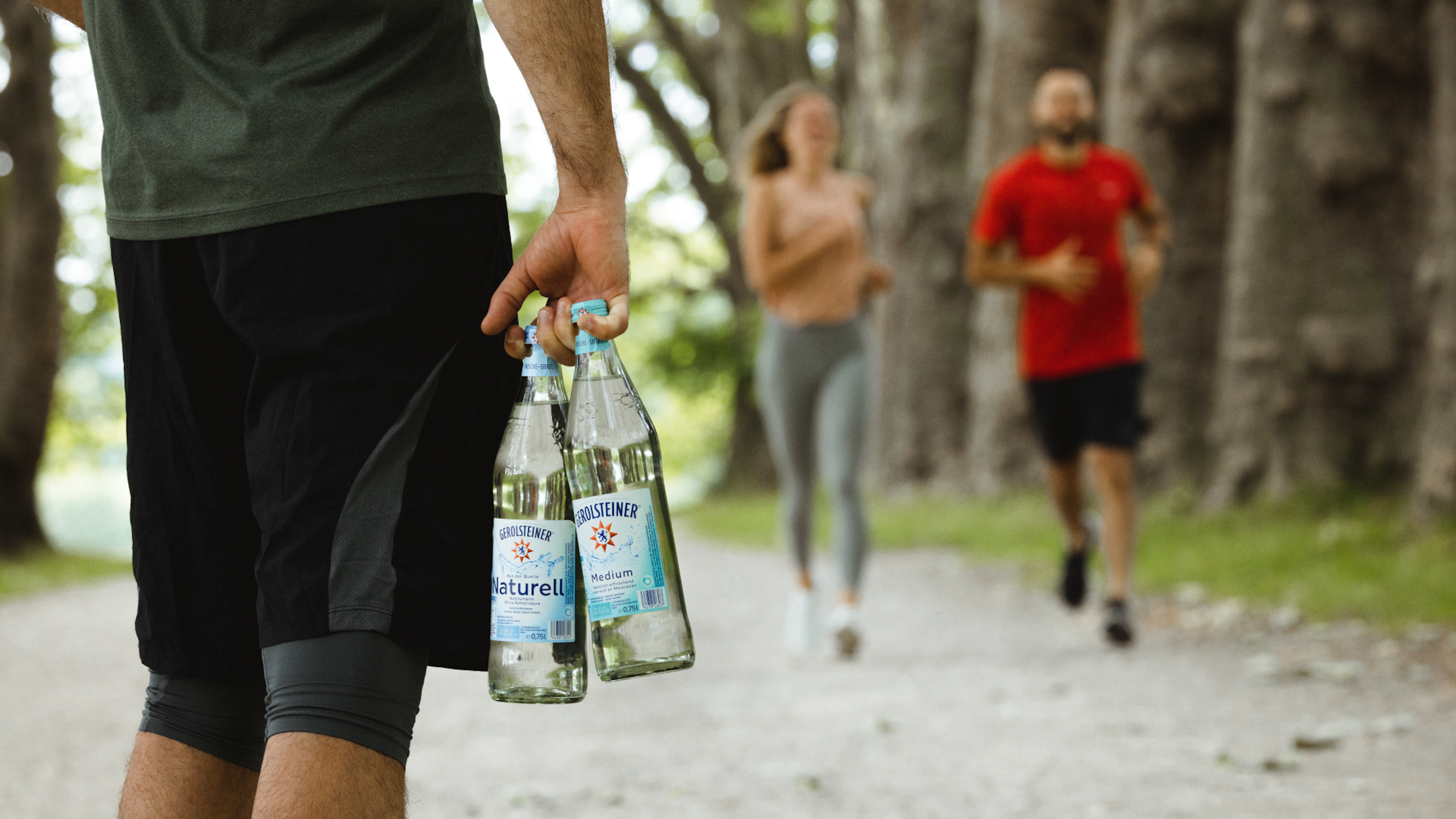 Ein Mann hält zwei Flaschen Mineralwasser, während im Hintergrund Personen joggen.